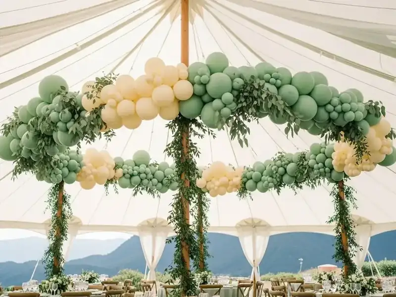 Guirnalda de globos verde salvia y crema con eucalipto tendida bajo el techo de una carpa de boda con vista al lago
