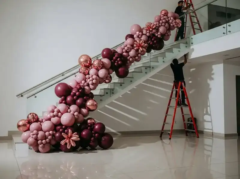 Guirnalda de globos color vino, rosa y rose gold cayendo por escalera moderna de barandal de cristal, equipo instalando